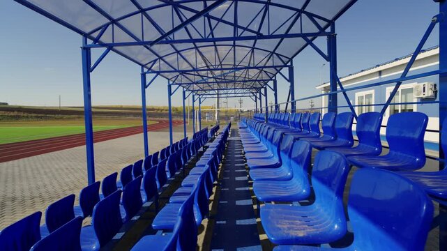Active view reveals row of plastic seats overlooking football field and running track. Sunlight illuminates empty stand hiding under canopy