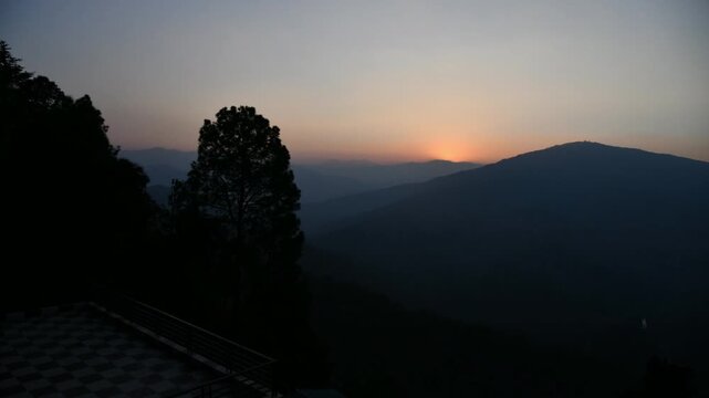 A scenic wide-angle shot of a golden sun setting behind layered mountain ranges in a tranquil hill station. The sky glows with a warm orange tint as the sun dips below the horizon.