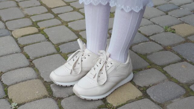 Close up of girl legs in white sneakers and high socks standing on pavement, A girl in white sneakers and lace-up knee-highs stands on the cobblestones
