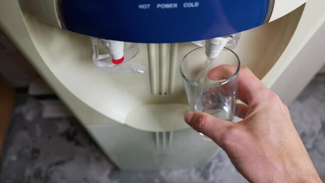A hand holds an empty clear glass cup under the cold dispensing tap of an indoor water cooler machine. Concept of daily hydration, healthy habits, and workplace refreshment breaks.