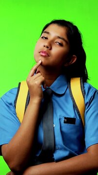 Portrait of Indian Asian teenage girl in school uniform with backpack standing against green chroma screen, looking thoughtful while placing hand on her chin, symbolizing curiosity, decision-making
