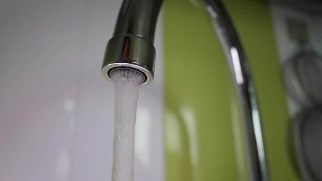 Aerated water flows steadily from a polished chrome kitchen tap in slow motion. Bright indoor lighting highlights the sparkling bubbles against a green and white background.