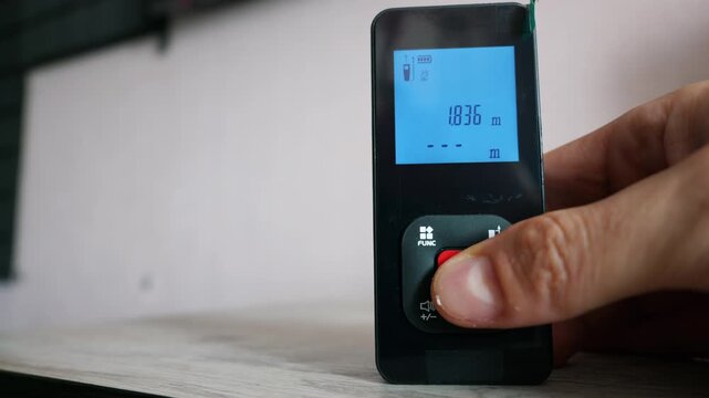 A hand holds a black digital laser distance meter over a light wooden surface in a bright room. The thumb presses a button on the device while the screen remains dark and reflective.