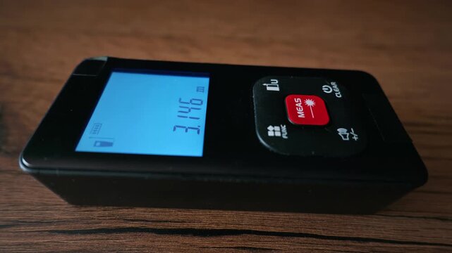 Finger presses the red button on a digital laser distance meter lying on a wooden surface. Concept of precision measurement, construction tools, and modern building technology.