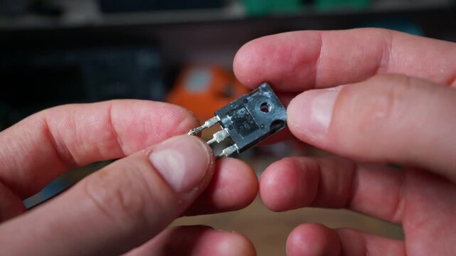 A close-up view of male hands holding and demonstrating a small electronic transistor component. The skilled engineer carefully examines the tiny microchip electrical element in a workshop.