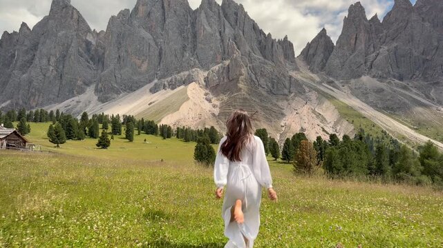 Beautiful Carefree Woman in White Dress Running Freely Through a Lush Alpine Wildflower Meadow Toward the Iconic Odle Geisler Spitzen Dolomite Peaks With a Traditional Alpine Hut, Val Gardena South Ty
