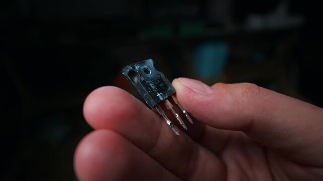 A person holds a power transistor electronic component with solder remnants on its metal pins. Concept of electronics repair, hardware engineering, microelectronics, and microchip assembly.