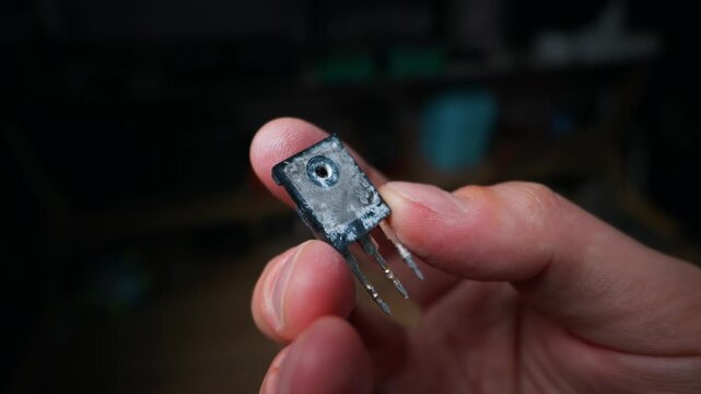 A close-up view of a male hand holding a dirty electronic power transistor component. The semiconductor device shows signs of wear and silver thermal paste residue on its metal heat sink.