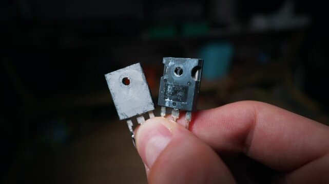 Two high power electronic transistors are held between the fingers of a hand. The close-up shot reveals technical markings and grey thermal grease on the metal backings of the hardware parts.