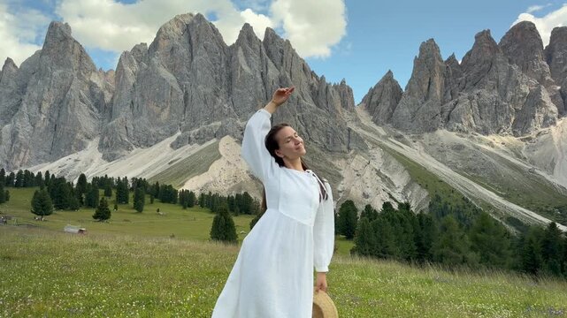 Happy Young Woman in White Dress Raising Hat and Smiling in Alpine Mountain Meadow, Odle Dolomites Italian Alps Val di Funes Italy