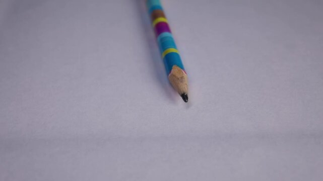 A close-up view shows a sharpened lead pencil with vibrant multi-colored stripes resting on a clean white sheet of paper. The soft lighting emphasizes the wooden texture and graphite tip.