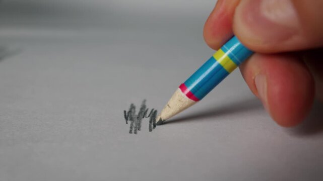 A close-up view shows a hand holding a colorful pencil and drawing on white paper. The graphite tip creates dark marks while being pressed against the clean surface under bright light.