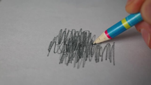 A human hand holds a blue and yellow pencil while scribbling a dark graphite patch on white paper. The close-up view focuses on the carbon layers being applied for a scientific experiment.