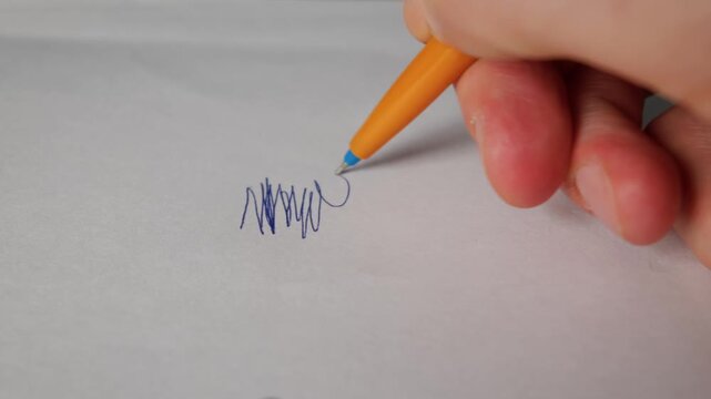 A hand scribbles blue ink zigzag lines with an orange ballpoint pen on a blank white sheet of paper. Concept of writer block, anxiety, testing a pen, and brainstorming ideas.