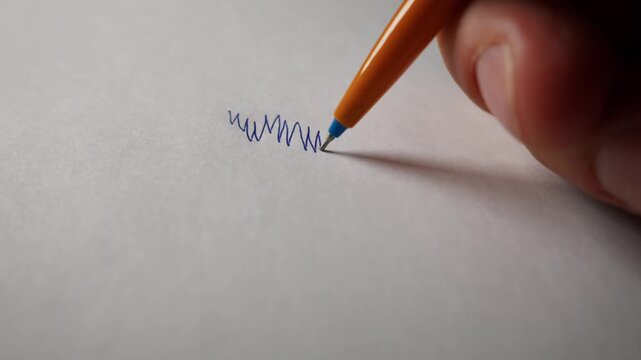 A hand holds an orange pen and scribbles blue ink lines on a white sheet of paper in a close-up shot. Concept of creativity, brainstorming, boredom, and testing office supplies.