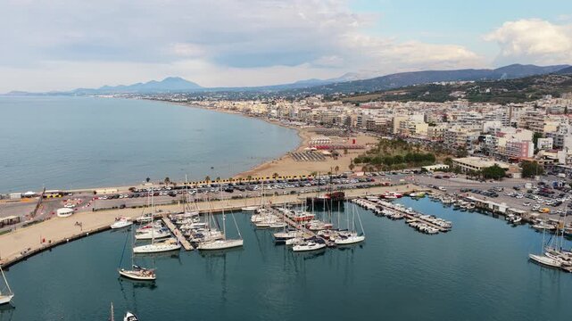 Aerial drone view of the marina and curving sandy bay with the seafront town below distant mountains, Rethymno, Greece