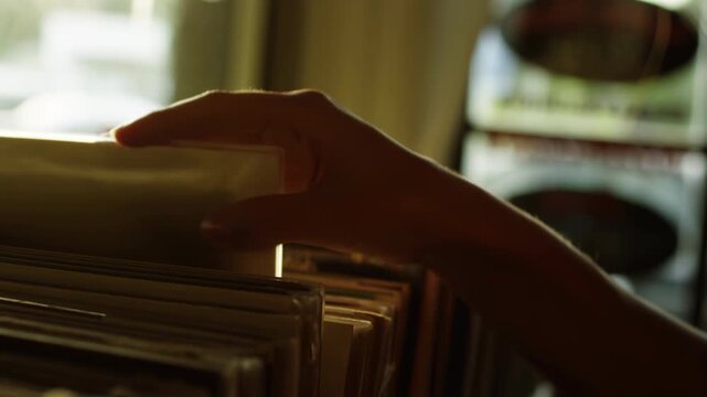 Hand pulling clear sleeve from stacked record rack under warm backlight with shallow depth of field and low-angle viewpoint, showing careful browsing, tactile selection, and subtle retail discovery