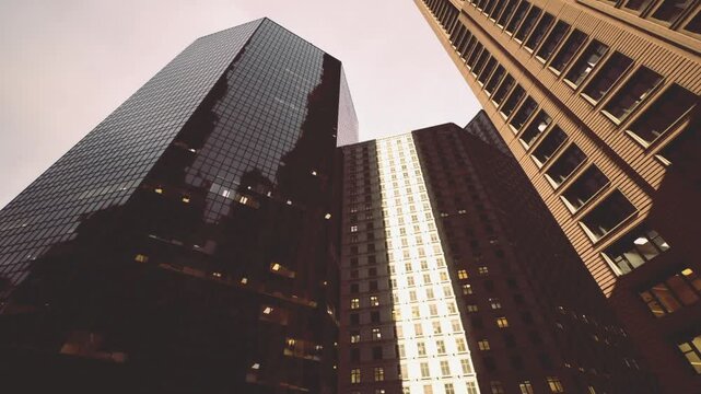 Dramatic upward view of converging towers framing small patch of sky, evening lights and textured surfaces create sense of depth and metropolitan intensity