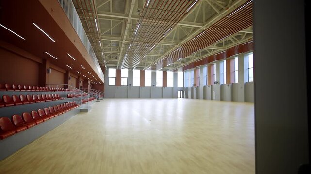 Dark doorway reveals massive sunlit sports arena with polished wooden floor. Ascending rows of orange stadium seats occupy left side