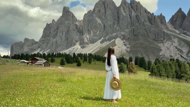 Beautiful Elegant Woman in White Dress and Straw Hat Facing the Majestic Odle Geisler Spitzen Dolomite Peaks in a Wildflower Alpine Meadow With a Traditional Alpine Hut, Val Gardena South Tyrol Italy