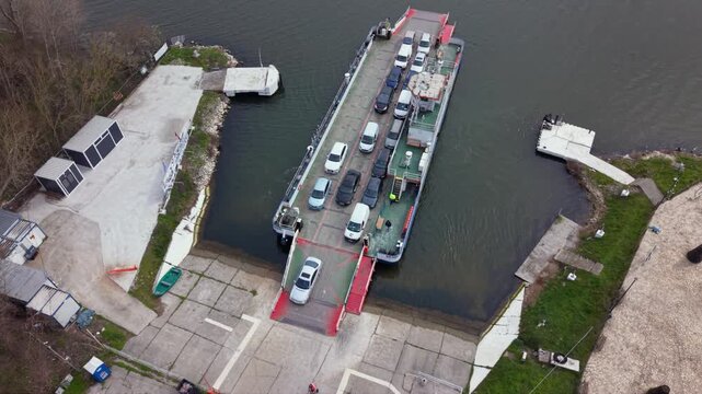 Aerial drone view of vehicle ferry transporting cars across a wide river with automobiles boarding and disembarking at ferry terminal connecting opposite riverbanks transportation infrastructure RO-RO