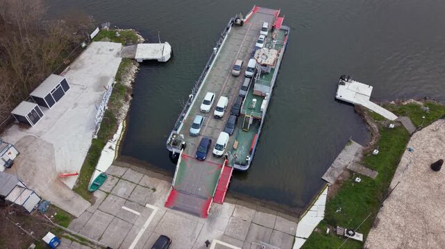 Aerial drone view of vehicle ferry transporting cars across a wide river with automobiles boarding and disembarking at ferry terminal connecting opposite riverbanks transportation infrastructure RO-RO