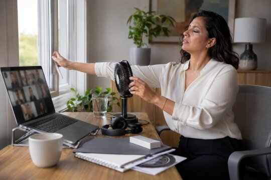 woman manages a hot flash during a remote work meeting, opening a window and using a small fan while maintaining a professional presence through cancer hormone therapy