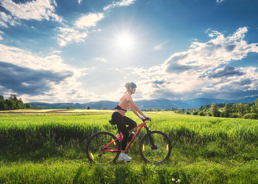 Female cyclist riding mountain bike through vibrant green meadow in Slovenia in summer. Woman cycling in scenic countryside landscape under bright sunny sky with lens flare. Active lifestyle. Sport