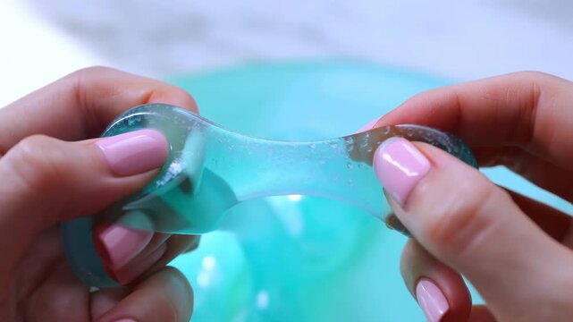 Hands stretching translucent teal slime with pale pink manicure, forming elastic bridge, glossy highlights, soft tension lines, rounded indentations, tiny bubbles, tactile close-up