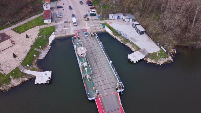 Aerial drone view of vehicle ferry transporting cars across a wide river with automobiles boarding and disembarking at ferry terminal connecting opposite riverbanks transportation infrastructure RO-RO