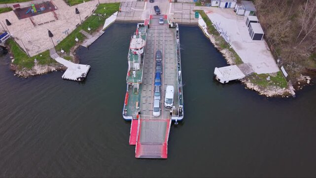 Aerial drone view of vehicle ferry transporting cars across a wide river with automobiles boarding and disembarking at ferry terminal connecting opposite riverbanks transportation infrastructure RO-RO