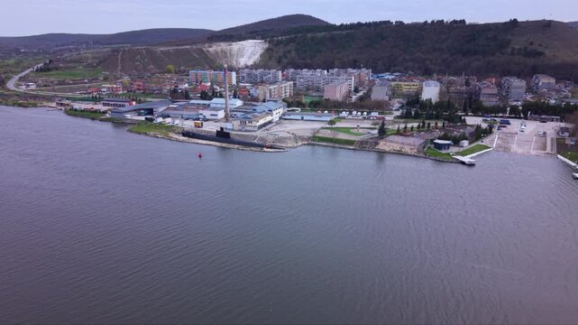 Aerial view of a submarine museum display beside a river, with the black submarine moored in a narrow channel near industrial buildings and rock-lined banks