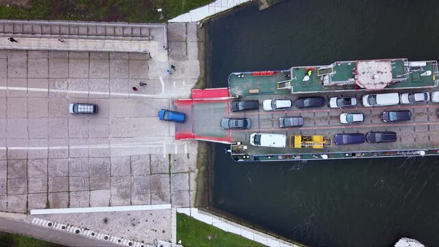Aerial drone view of vehicle ferry transporting cars across a wide river with automobiles boarding and disembarking at ferry terminal connecting opposite riverbanks transportation infrastructure RO-RO