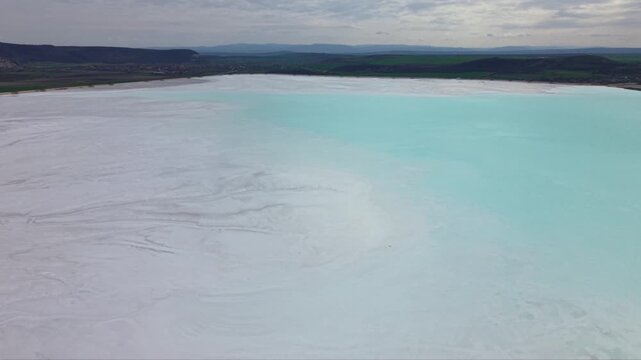 Aerial drone footage flying low above white mineral evaporation pond with intricate natural patterns and textured sediment surface in industrial extraction area surrounded by hills dramatic cloudy sky