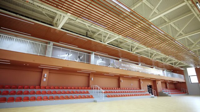 Rows of empty plastic chairs fill hall showing scale of sports venue. Massive fencer murals cover wall sections hanging above railing