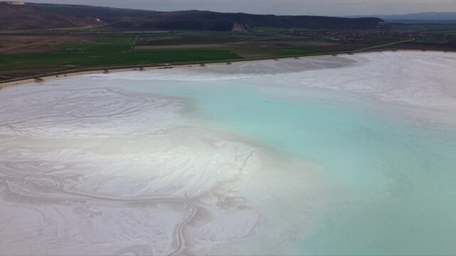 Aerial drone footage flying low above white mineral evaporation pond with intricate natural patterns and textured sediment surface in industrial extraction area surrounded by hills dramatic cloudy sky