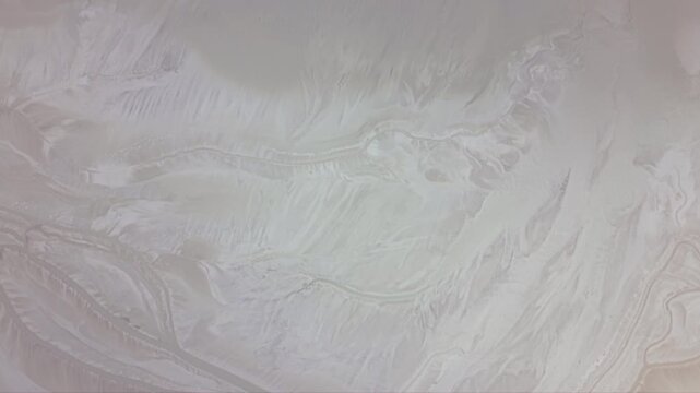 Aerial drone footage flying low above white mineral evaporation pond with intricate natural patterns and textured sediment surface in industrial extraction area surrounded by hills dramatic cloudy sky