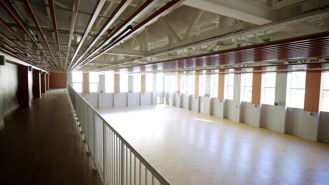 Upper level walkway with dark floor and metal handrail overlooks empty sports hall. Large windows provides natural daylight along wall