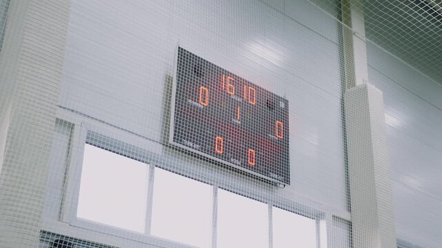 Electronic digital scoreboard glows behind protective safety net in sports hall. Active timing display tracks professional match statistics