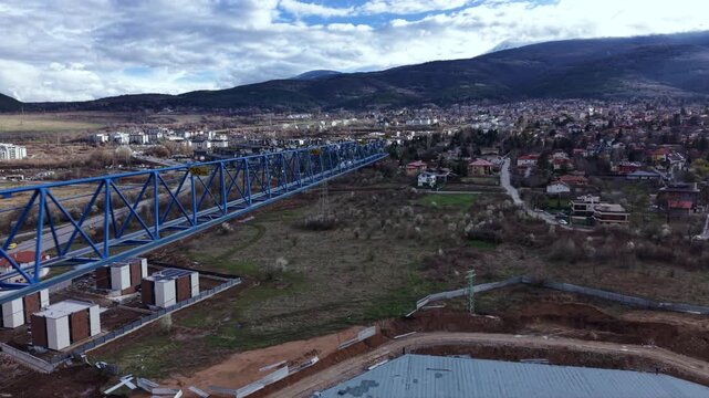 Aerial pass beside the long blue lattice jib of a tower crane above a construction site, with suburban houses and a mountain beneath heavy clouds.