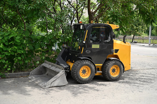 Yellow skid steer loader parked on an asphalt road near green trees for city maintenance.