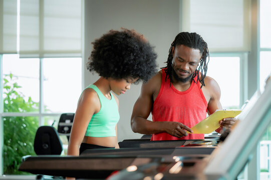 Young girl with curly hair exercises seriously on treadmill. Man beside her watches and guides her. Both focused and determined to improve fitness. Ideal for workout, coaching