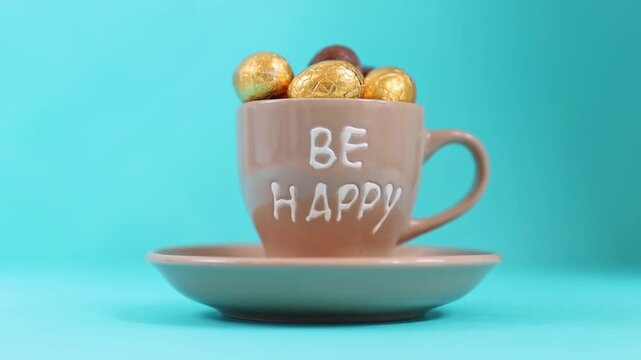 One coffee cup with the inscription Be Happy in a saucer full of a bunch of chocolate mini egg candies in a gold shiny wrapper, lies on a blue background with changing soft light and shadow, side view