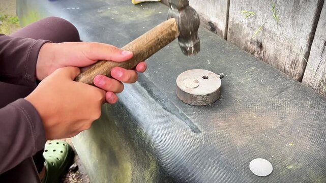 A close-up view of a person using a vintage hammer to flatten a circular metal blank on a small round steel anvil outdoors.