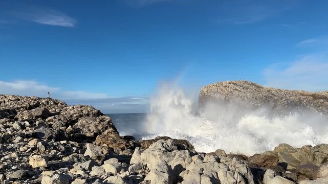  Close-up of powerful Atlantic waves crashing against coastal rocks, sending white foam toward the camera. Sunlit sea spray creates a rainbow beneath a clear blue sky with authentic ocean wave sound.