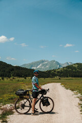 Fototapeta premium Man standing with a loaded gravel bicycle in Durmitor National Park, Montenegro. Summer bikepacking adventure surrounded by alpine mountain scenery.