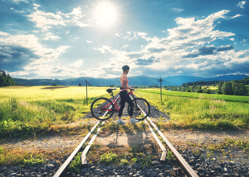 Female cyclist riding mountain bike on gravel country road in Slovenia. Woman cycling next to green fields and countryside railway station under blue sky with sun, clouds. Active lifestyle. Railroad