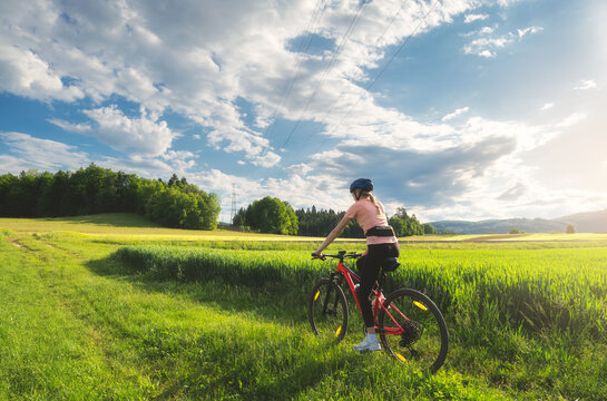 Female cyclist riding mountain bike on gravel path through green fields in Slovenia in summer. Woman cycling away into scenic countryside landscape at sunset. Active lifestyle. Girl on bike. Sport