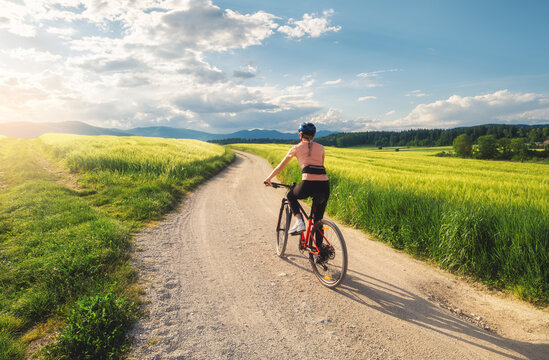 Woman riding mountain bike on dirt road through green wheat fields at sunset in Slovenia. Female cyclist cycling in countryside landscape and sunny sky. Summer outdoor activity, fitness. Girl on bike