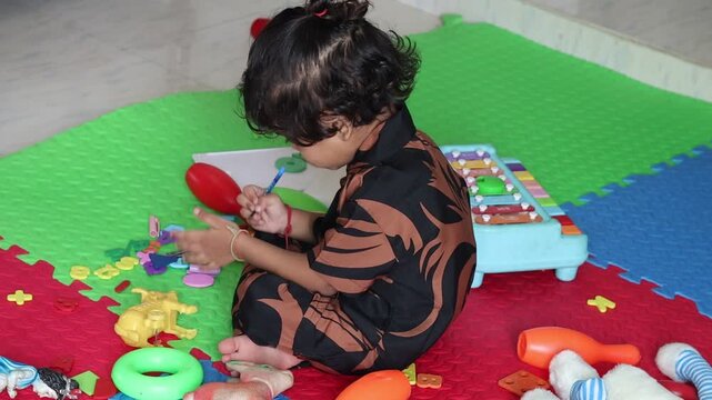 Side profile of a cute Indian toddler girl sitting on a colorful playmat surrounded by scattered educational toys, holding a pen and playfully drawing on her own hand indoors.
